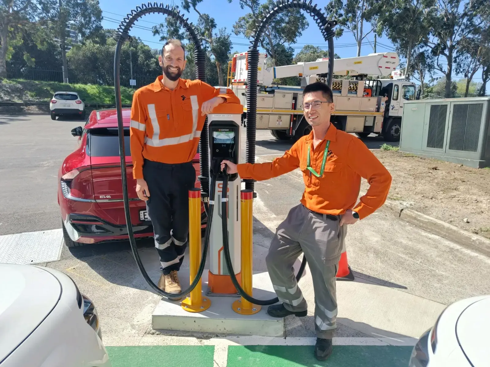 Two field technicians beside a dual-cable DC fast charger managed by LinkRay, with an EV parked behind